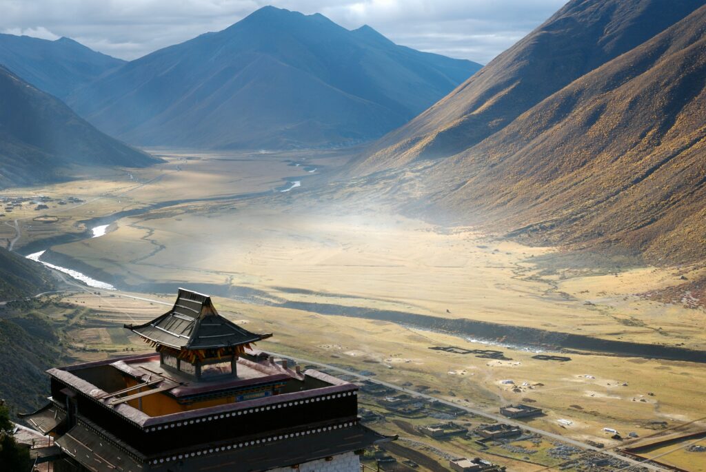 A breathtaking view of a traditional Tibetan monastery perched on a hill, overlooking a vast valley surrounded by towering mountains