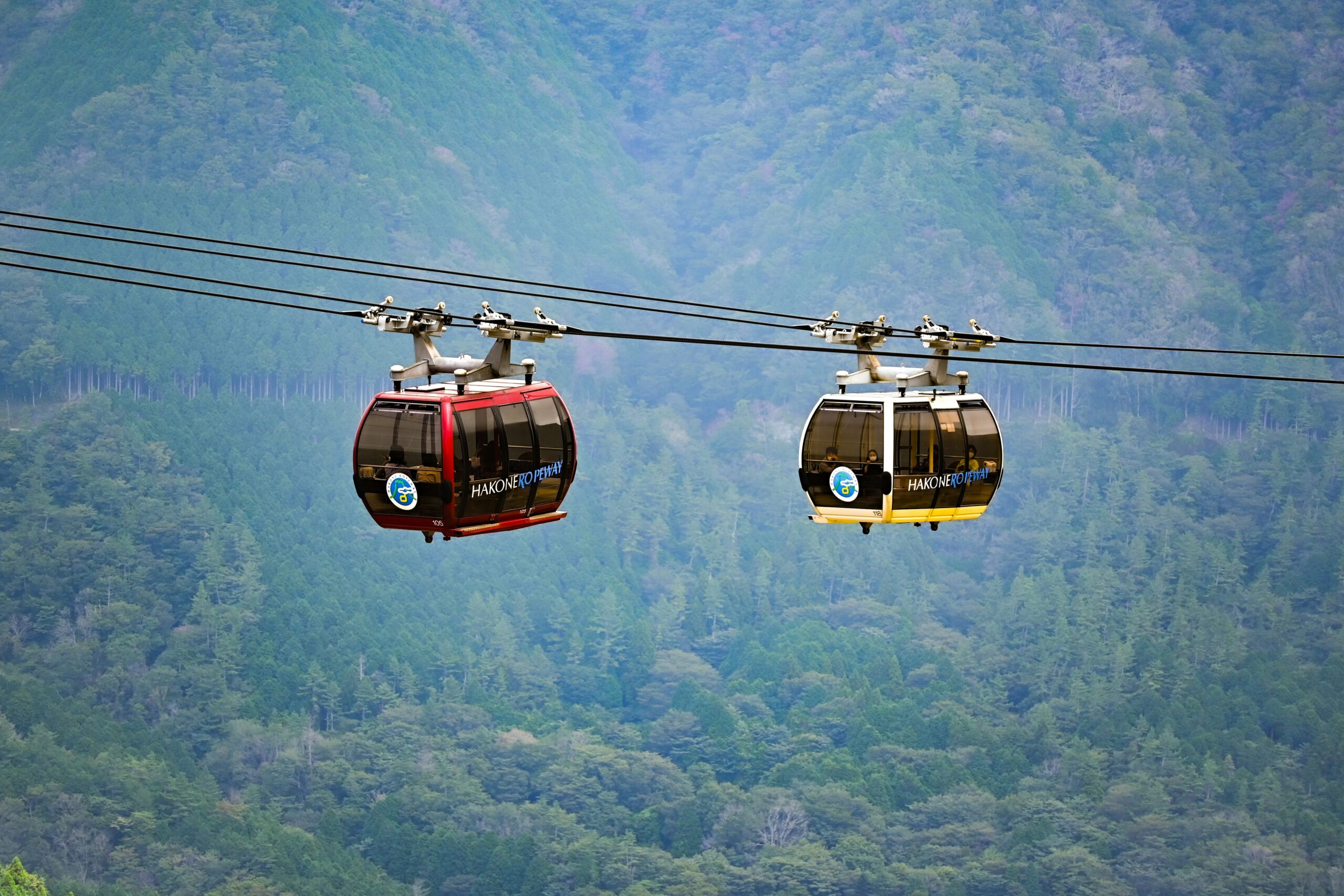 Two gondolas of the Hakone Ropeway, one red and one yellow, suspended in mid-air against a lush green mountainous background