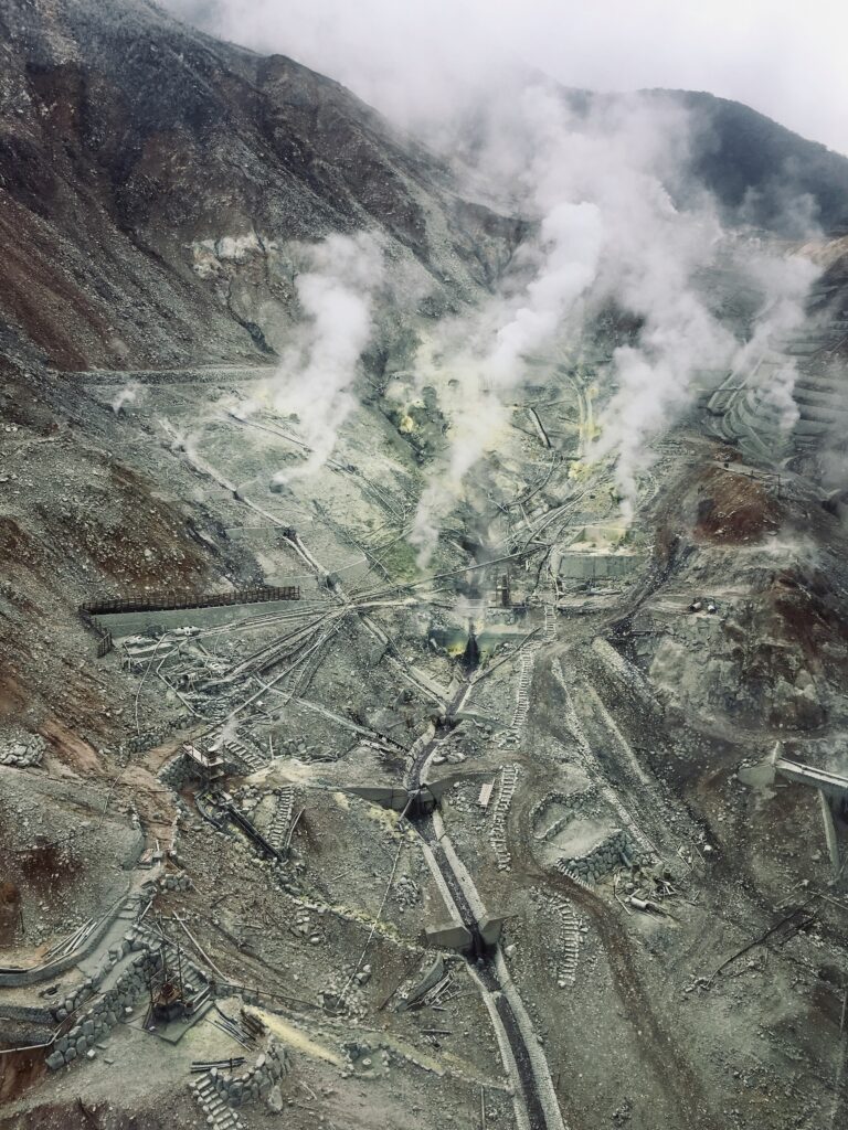 An aerial view of a rugged volcanic landscape with steam rising from multiple geothermal vents