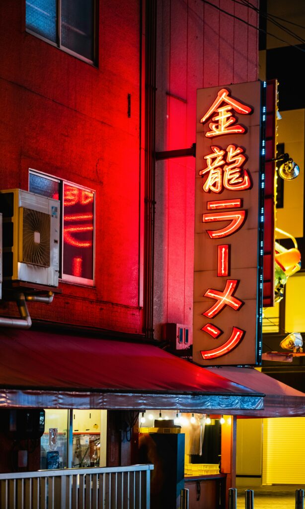 A nighttime street view of a traditional Japanese ramen shop with a glowing red neon sign that reads "ラーメン" (ramen)