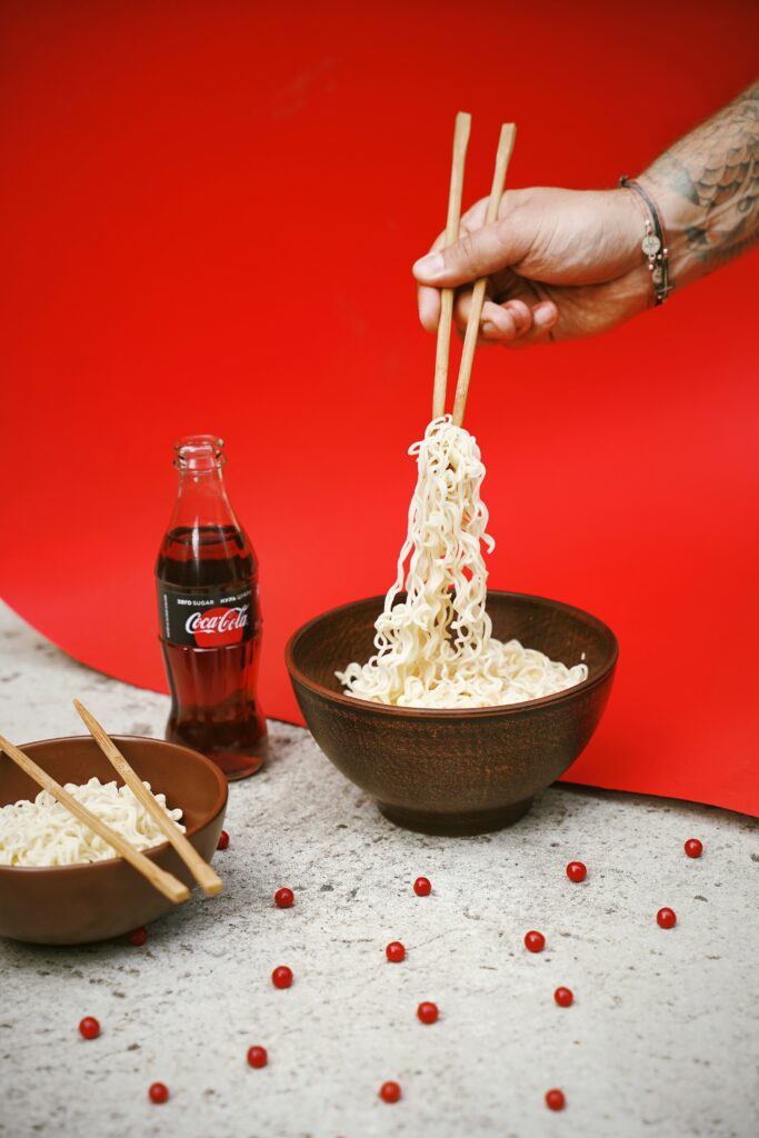 A minimalist and modern food composition featuring a hand with tattoos using chopsticks to lift instant noodles from a dark wooden bowl